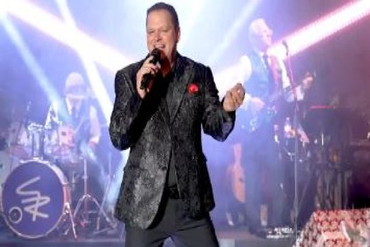 A man in a patterned suit sings into a microphone on stage while a band performs in the background with colorful lights.