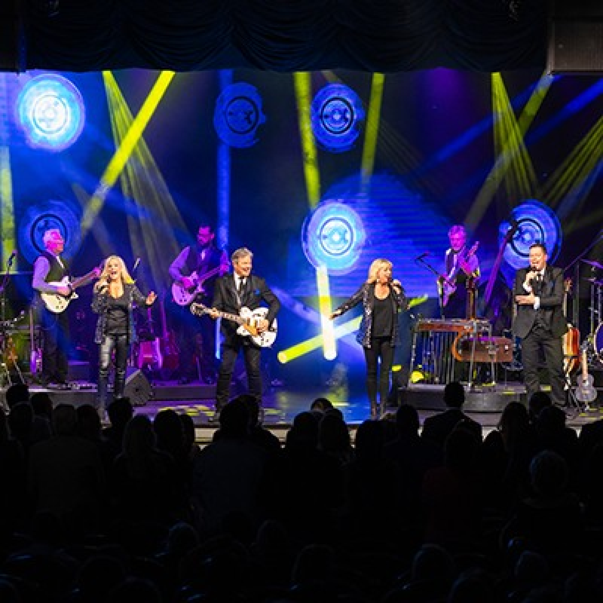 A band performs energetically on stage with bright lights and musical instruments, including guitars and drums, in front of an audience.