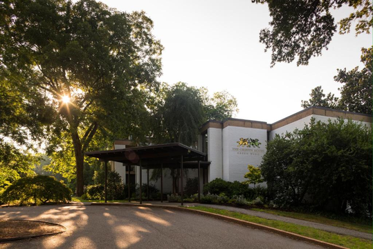 Sunlit exterior of a modern building surrounded by trees, featuring a sign with a horse logo on the facade.