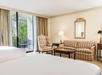 Hotel room interior with two beds, a striped sofa, armchair, coffee table, and a lamp. Sliding glass door leads to a small balcony with outdoor seating. Beige carpeting and curtains.
