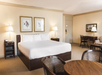 A hotel room with a king-size bed, two bedside lamps, a desk, a chair, and a round table with chairs. The walls feature framed artwork.