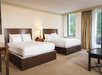 Hotel room with two double beds, a nightstand with a lamp between them, a window with curtains, and a corner table with chairs.