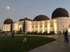 Tourists standing and sitting in front of the front of Griffith Observatory at dusk with the moon shining over head.