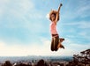 A woman with very curly hair wearing a pink tank top jumping in the air with her arms up on sunny day.