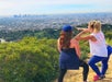 Two women in athletic wear facing away from the camera pointing out something in the valley below them with Los Angeles in the background.