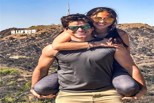 A man and woman wearing sunglasses with the woman on the mans back smiling with the Hollywood Sign in the background.