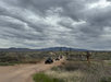 Four people ride ATVs on a desert dirt path under a cloudy sky, surrounded by sparse vegetation and distant mountains.
