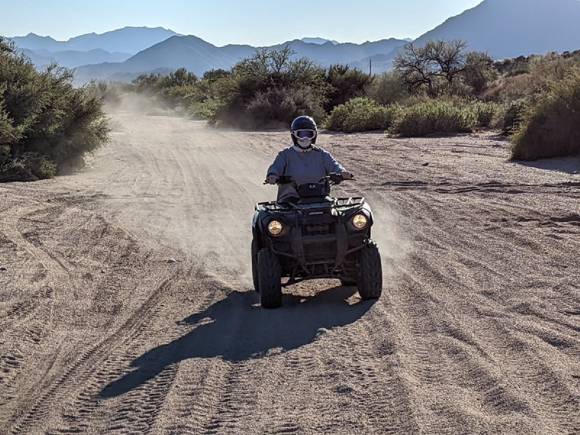 Person riding an ATV on a dusty trail in a desert landscape with mountain backdrop and clear sky.