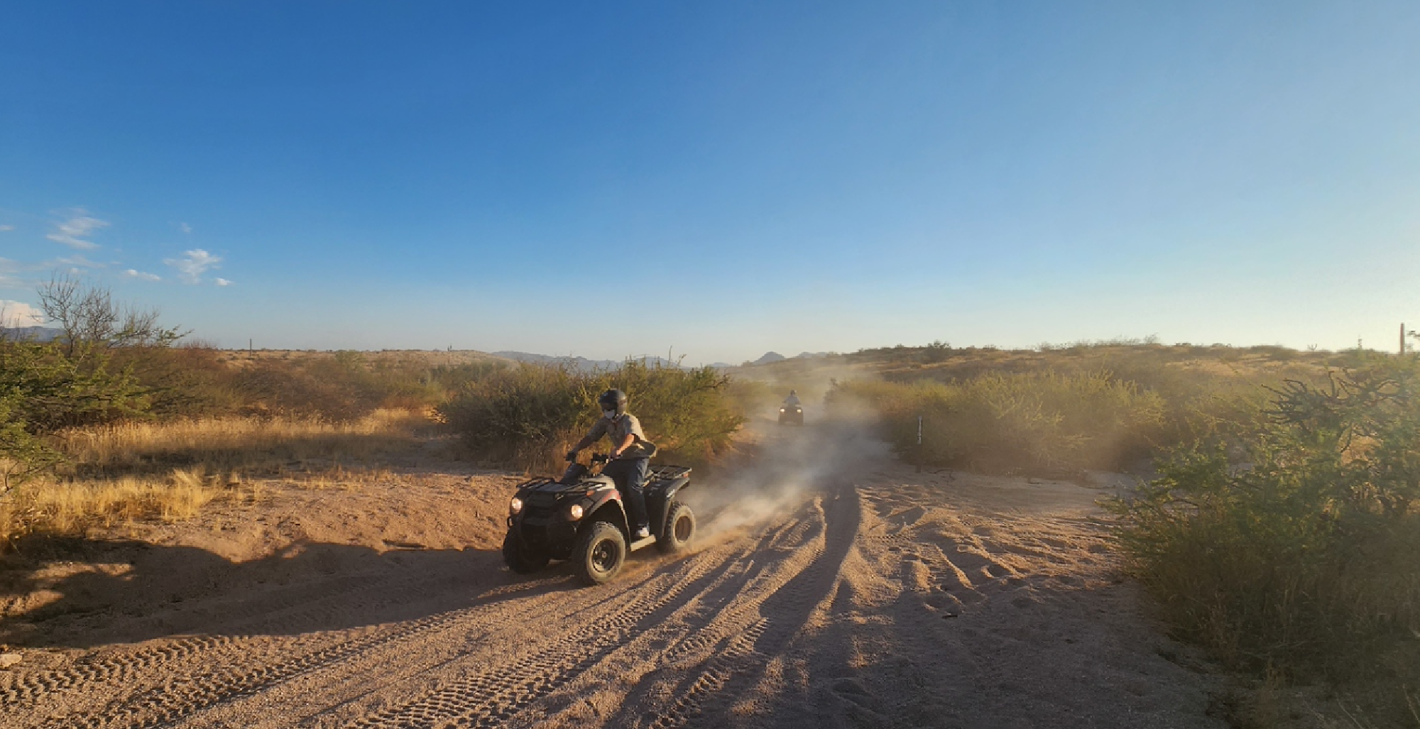 Person riding an ATV on a sandy trail in a desert setting under a clear blue sky, with dust trailing behind. Sparse vegetation is visible in the background.