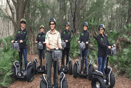 Group of tourists wearing black jackets on segways in the forrest with their tour guide wearing a khaki shirt on a segway in front of them.