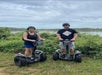 A man and woman wearing helmets and standing on segways with bushes and water behind them on the Guided Segway Tour of Lake Louisa State Park.
