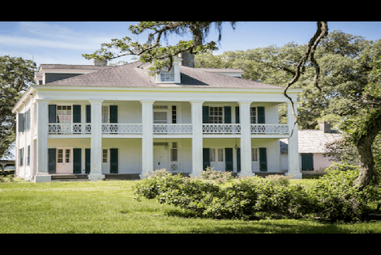 Large two-story white plantation house with tall columns, green shutters, and a balcony, surrounded by grass and trees under a partly cloudy sky.