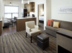 A modern hotel room with a living area with a couch and chair and a kitchenette with a dining table and a bedroom visible through a doorway.