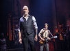 A man in a pinstripe suit stands on stage with a serious expression, while another person plays a guitar in the background. Stage lights cast dramatic shadows.