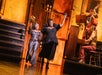 Two women stand on stage, one holding flowers and the other raising a hat, while two musicians play piano and upright bass in the background.