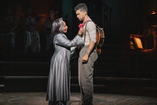 A woman in a long coat pins a red flower to the shirt of a man with a guitar on his back, both standing on a dimly lit stage.