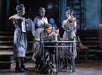 A young woman sits at a desk reading a letter, surrounded by three women in patterned dresses, one with an accordion and another playing a violin, on a dimly lit stage.