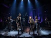 Performers on stage in dramatic lighting and costumes, surrounded by fog, during a theatrical production.