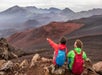 Two people with backpacks sit on rocky terrain, one pointing towards distant mountains under a cloudy sky.