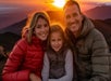 Three people, two adults and a child, sit close together outdoors wearing jackets, smiling at the camera with a sunset and mountains in the background.