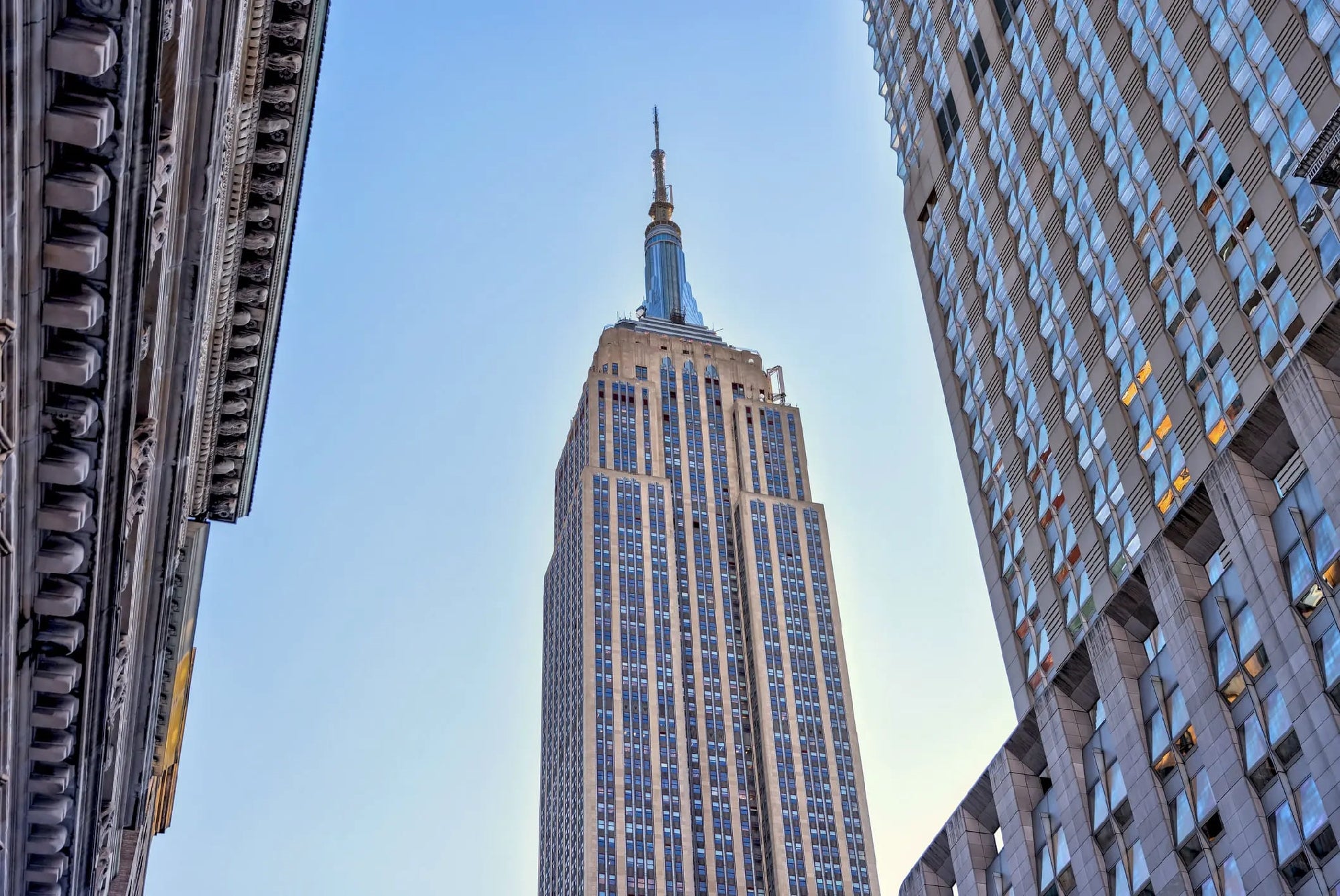 The image shows the Empire State Building in New York City, viewed from below with adjacent buildings framing it and a clear blue sky in the background.