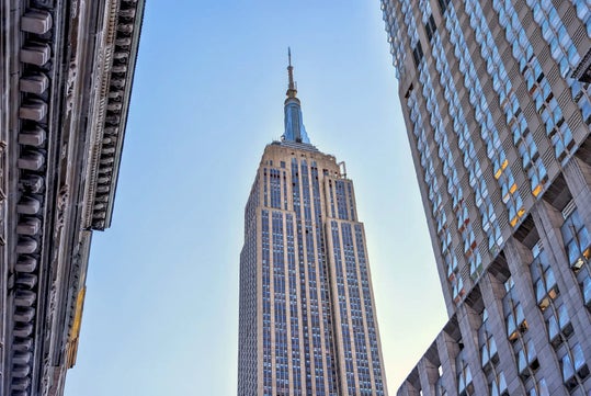 The image shows the Empire State Building in New York City, viewed from below with adjacent buildings framing it and a clear blue sky in the background.