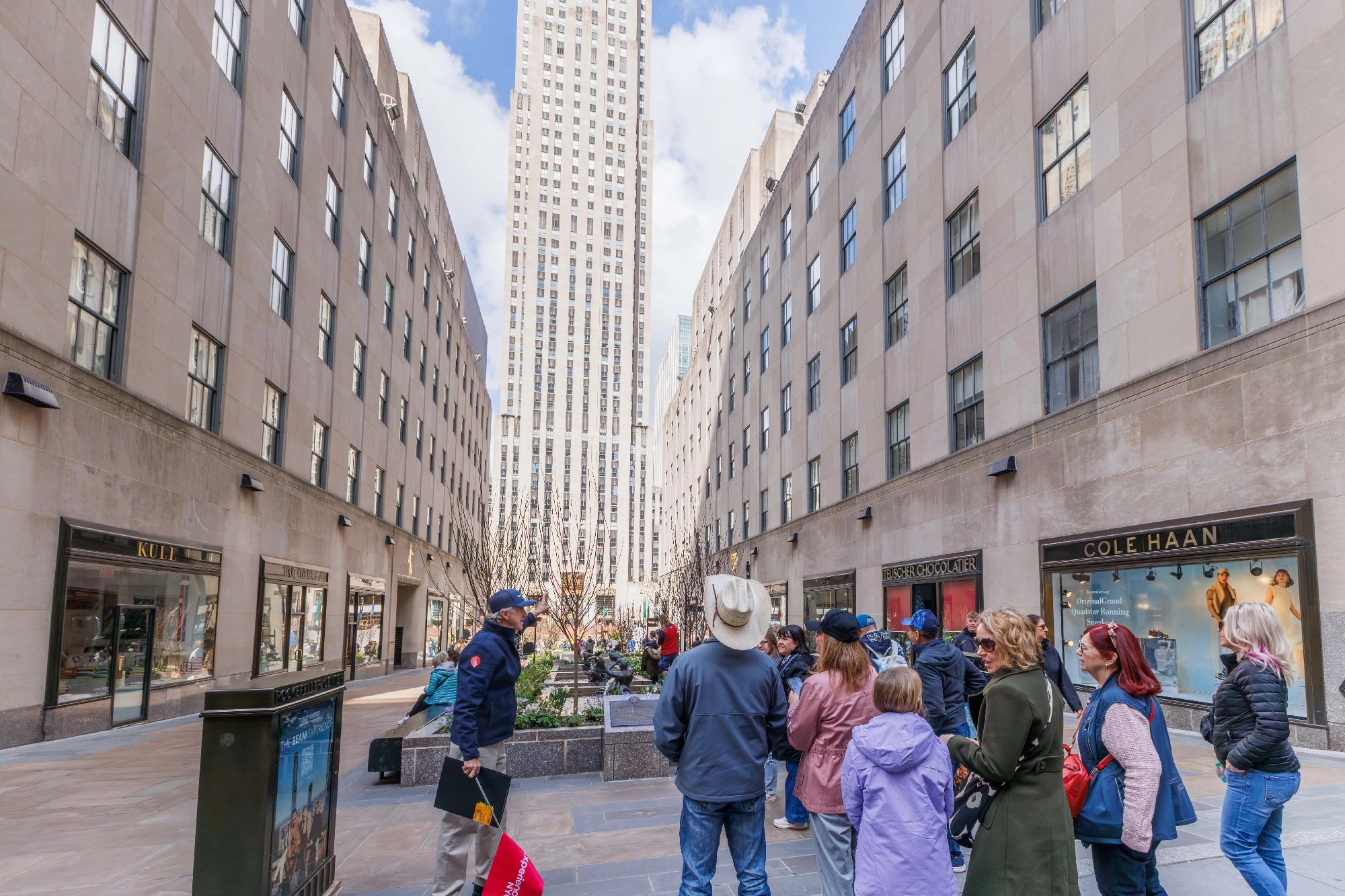 A group of people listen to a guide in an outdoor plaza between tall buildings with retail shops, under a partly cloudy sky.