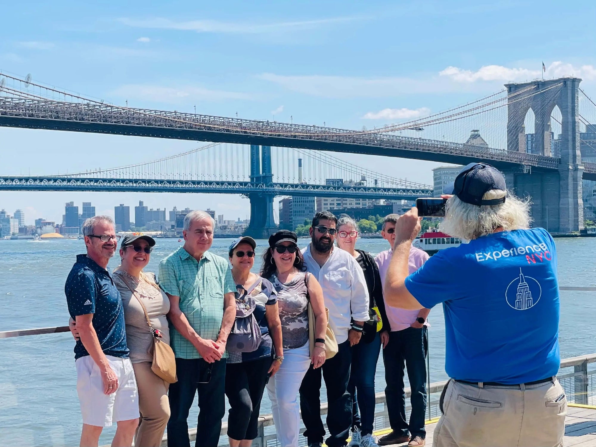 A group of eight people pose for a photo near the waterfront with the Brooklyn and Manhattan Bridges visible in the background on a sunny day.