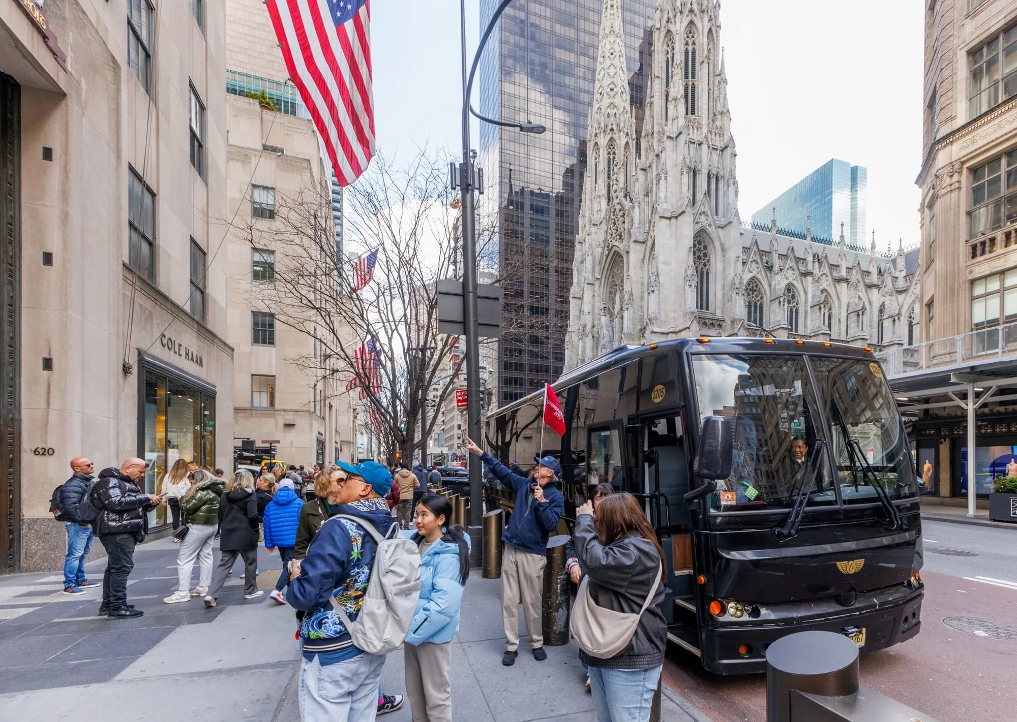 A group of people gathers near a black tour bus parked on a city street with tall buildings and an American flag visible.