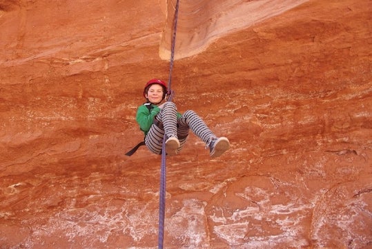 A child in a green jacket and red helmet sliding down a rock climbing rope with red rock behind her on the Half-Day Moab Canyoneering Adventure.