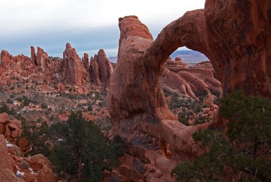 Stunning single arch rock formation at the Arches National Park with foliage scattered everywhere and a cloudy sky overhead.