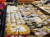 Cheese display in a market with various types and brands, arranged in rows, including rounds, wedges, and blocks. Labels with names and prices are visible on some products.