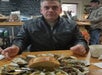 Person sitting at a table with a plate of oysters and bread, wearing a leather jacket. Another person in the background near a menu board.