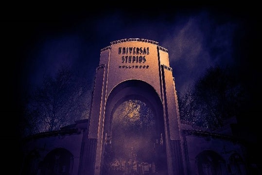 The concrete arch front entrance of Universal Studios Hollywood with dark purple fog around it and spooky leafless trees in the background.