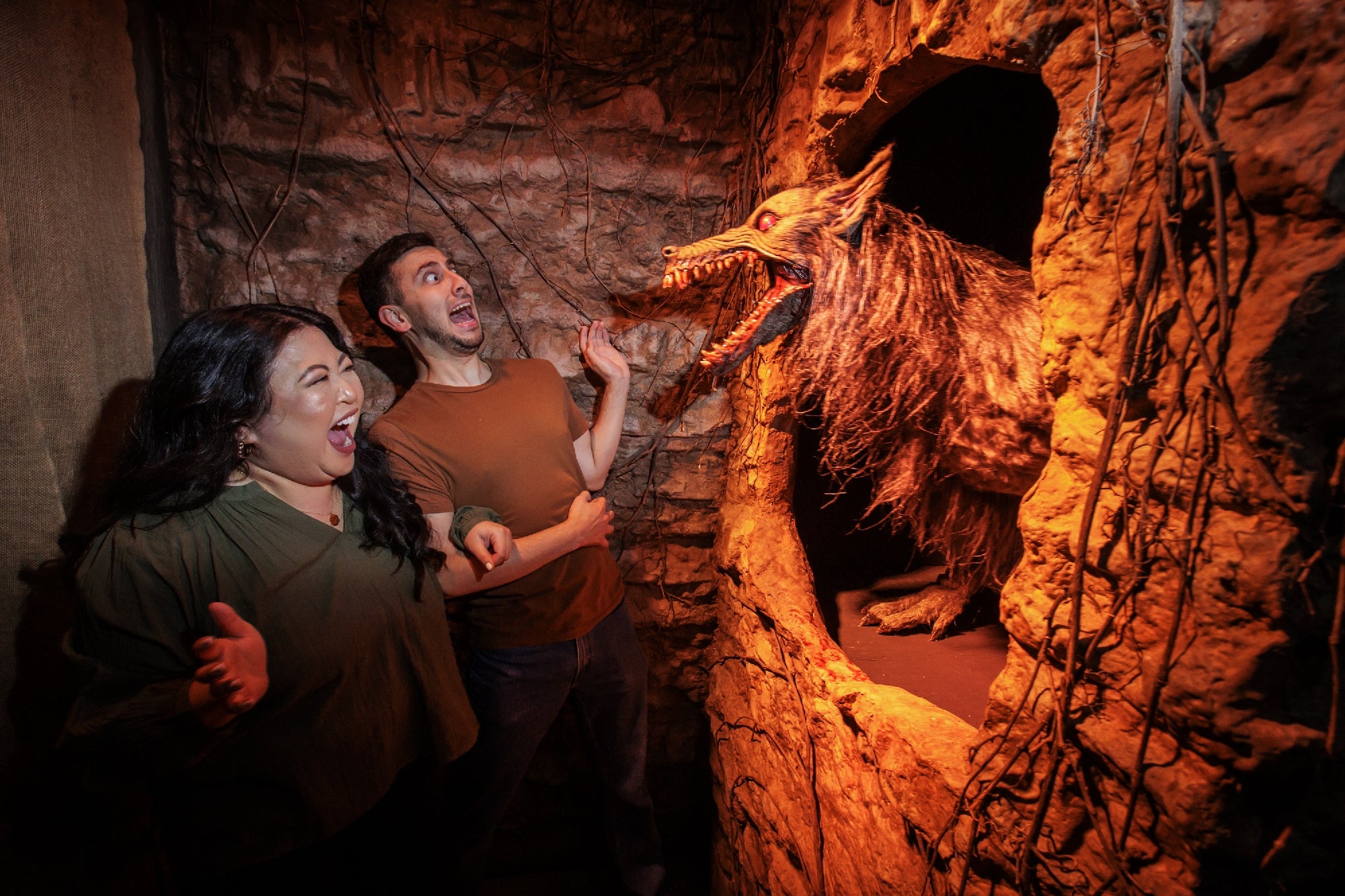 Two people react with shock and fear as a large, snarling wolf-like creature lunges from a hole in a rocky wall in a dimly lit setting.