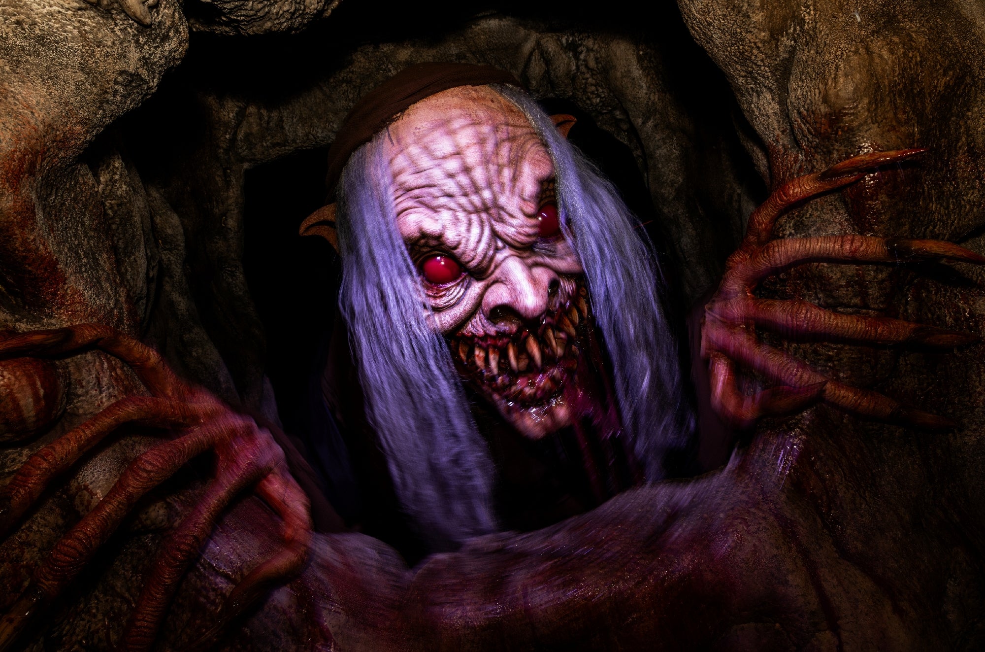 A monstrous creature with red eyes, long white hair, sharp teeth, and large claws emerges from a dark, rocky hole.