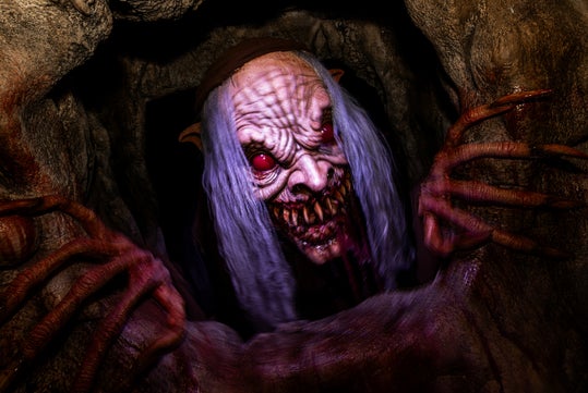 A monstrous creature with red eyes, long white hair, sharp teeth, and large claws emerges from a dark, rocky hole.