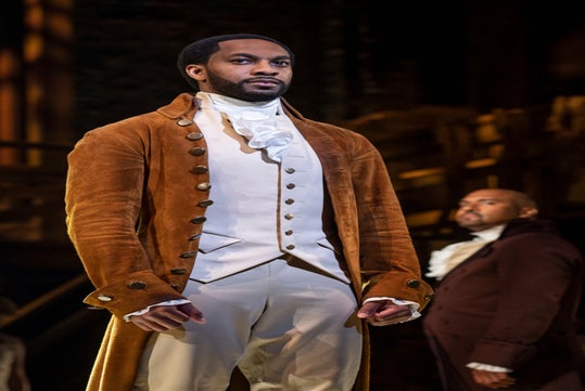A man in historical attire stands confidently on stage, wearing a brown coat, white vest, and cravat, with another man in similar costume blurred in the background.