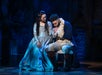 Two actors in period costumes perform a dramatic scene on stage, illuminated by blue lighting.