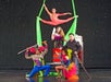 Five circus performers pose on stage: an aerialist in a split above, a woman holding a dove, and three others in colorful costumes holding swords in front of a black background.