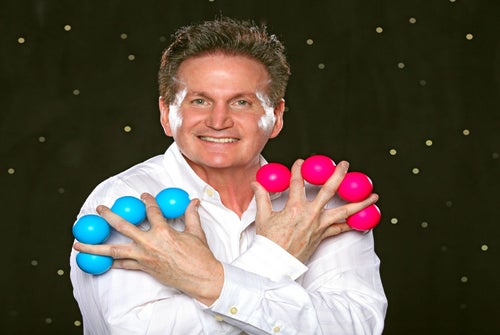 A man in a white shirt smiles, holding blue and pink balls between his fingers, with white makeup on his cheeks, against a black background with small dots.