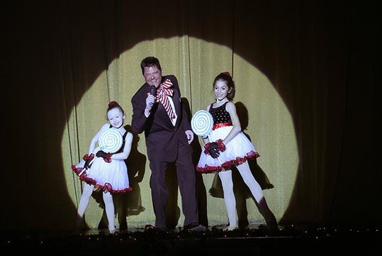 A man in a suit holding a microphone stands between two young girls in festive dance costumes holding large lollipops, all under a spotlight on stage.