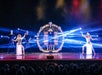 A magician stands inside a large circular prop onstage, flanked by two dancers posing, with bright stage lights and two assistants standing in the background.