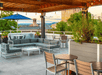 A rooftop patio with a lounge area and several tables with potted plants around them and the city in the background.