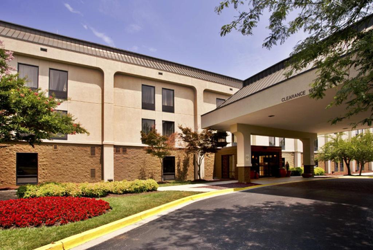 Hotel exterior with entrance canopy, surrounded by landscaping, trees, and a red flower bed.