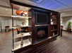 Modern lounge area with a dark wood entertainment unit featuring a black TV screen, a decorative fireplace, and various shelves. There are tables and chairs in the background.
