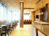 Bright, modern dining area with a row of tables and chairs on the left, a central column, and high chairs at a counter. Pendant lights hang from the ceiling.