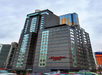 Tall, modern Hampton Inn hotel building with glass windows and red signage, surrounded by cityscape and traffic. Cloudy sky overhead.