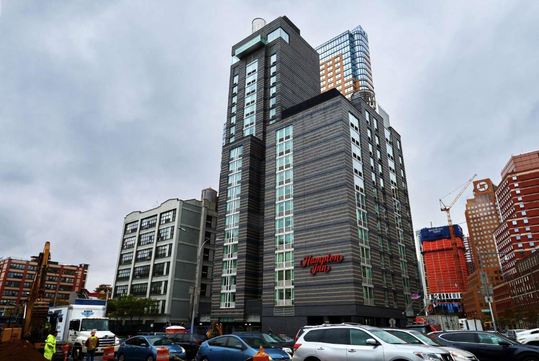 Tall, modern hotel building with "Hampton Inn" sign, surrounded by construction and parked cars under a cloudy sky.