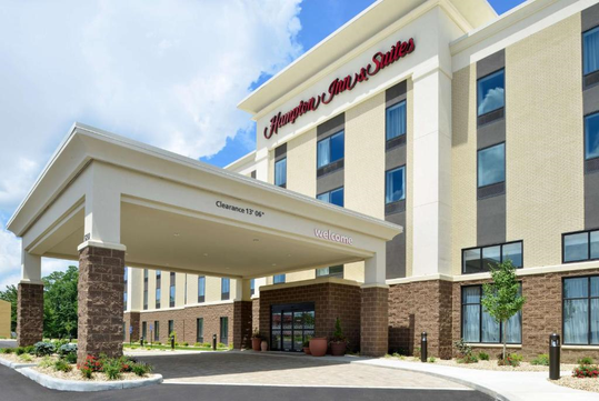 Exterior of a Hampton Inn & Suites hotel with a covered entrance and a "Welcome" sign. The building has a beige and brown facade with multiple windows.